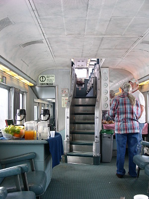 [A slightly curved stairwell with seven steps is in the middle of the car and has an opening to the upper level. A table with fruit and drinks sits on the left side of the car. Two people standing and talking are on the right side of the car.]